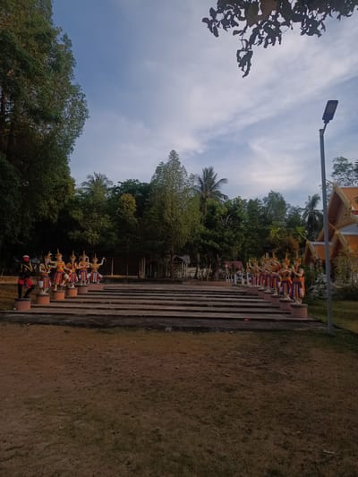 The image depicts a row of statues on concrete pedestals lining a set of concrete stairs, situated in Baan Wok Tum, Thailand. The statues appear to be figures from Thai mythology or folklore, adorned with colorful garments and headdresses. The stairs lead towards a dense area of trees and foliage, partially obscuring buildings in the background. To the right, a modern street lamp is visible next to traditional Thai architectural elements with gilded details. The foreground features a dry, grassy area. The sky above is lightly clouded, suggesting either dawn or dusk. There are no people visible in the frame other than the statues.