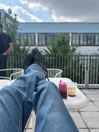 The image captures a first-person perspective of someone relaxing outdoors on a patio or balcony in Turin, Italy. The viewer's legs, clad in blue jeans and black shoes, are crossed and rest on what appears to be a white outdoor chair. To the right, on a white surface (likely part of the chair or a small table), sits a clear plastic cup containing a yellowish liquid, possibly a beverage. Next to the cup is a pink electronic device, identified by the visible text as "WATERMELON ICE" and "Elfbar 2nd."
In the middle ground, a metal railing separates the patio from the surroundings, which include green trees and other buildings. A modern-looking building with horizontal light blue panels and dark-framed windows dominates the background. Above, the sky is partly cloudy and bright, with sunlight breaking through.
In the lower left corner, a portion of another person is visible, wearing a dark shirt and possibly a cap, with their right arm resting on their hip. Further in the background on the left, another building with a satellite dish is visible. The floor of the patio appears to be paved with light-colored tiles. The scene conveys a moment of leisure during the daytime, likely under pleasant weather conditions in an urban setting.