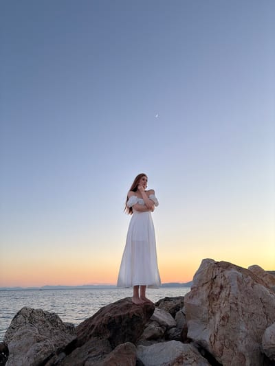 A young woman stands on large rocks near the sea in Paleo Faliro, Greece, at dusk. She is wearing a white, off-the-shoulder dress. The sky is a gradient of blue, transitioning to orange and yellow near the horizon. A crescent moon is visible in the upper portion of the sky. The sea is calm, with a distant coastline visible. The rocks are varied in size and texture, with some showing signs of erosion.