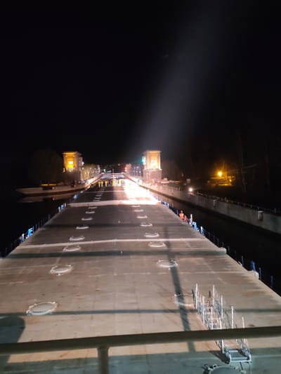 The image depicts an elevated, concrete structure at night, possibly a dam or large canal feature, illuminated by lights. The structure has a series of circular access points along its surface. Two tower-like structures with illuminated windows are visible in the mid-distance, with a line of lights stretching between them, suggesting a road or pathway. People are visible in the distance between the towers. One individual is sitting on the railing to the right of the structure. Trees and vegetation are present along the sides, and water is visible on both sides of the structure. The location is Водник Снт, Russia.