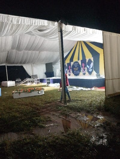 This image captures an artificially lit outdoor event tent at night in Marenyo-Shianda ward, Kenya. The tent features a white, draped fabric ceiling and white side panels. The ground beneath the tent is wet and muddy, with numerous puddles reflecting the interior lighting, indicating recent precipitation.
A raised stage is positioned at the far end of the tent, adorned with a black and yellow radially striped backdrop. On the stage, three large vertical banners are displayed: the left and right banners feature portraits of women, while the central banner depicts a white dove. Below these images, partially legible text is present. Various pieces of event equipment, including dark-colored speakers or equipment boxes, are visible on and around the stage. To the left of the stage, stacked equipment racks are present, alongside a white cushioned bench.
In the foreground, a rectangular white planter box containing orange and white flowers is situated on the muddy grass. A single individual, wearing a red long-sleeved shirt, blue jeans, and light-colored boots, is partially visible to the right of a central tent support pole, walking towards the right side of the frame. The scene conveys an active event space, possibly a memorial or celebration, set up under challenging environmental conditions.