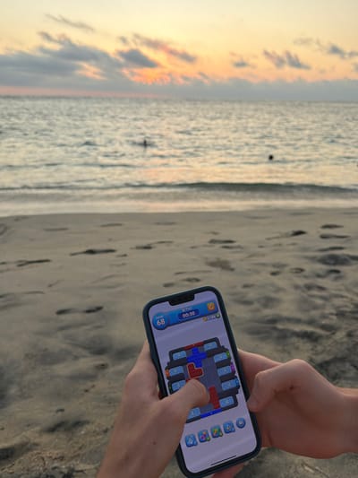 At a beach, someone holds an iPhone displaying a game with colorful blocks, engaging with their finger. The ocean and a vibrant sunset are blurred in the background. The sandy beach is visible beneath the person's hands and phone. The scene captures a moment of leisure and technological engagement against a natural backdrop.