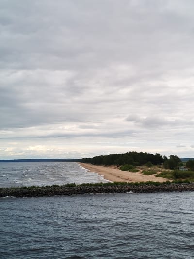 The image depicts a view of a coastline in Ozernoe Ust'e, Russia, on a cloudy day. In the foreground, dark blue water with visible ripples fills the bottom half of the frame. A breakwater made of dark rocks and green vegetation runs horizontally across the middle of the image, separating the foreground water from the calmer water beyond.
Beyond the breakwater, a sandy beach stretches along the coastline. A dense line of dark green pine trees borders the beach. Further back, the land appears to rise slightly, with more trees and a small, light-colored building with a dark roof visible amongst them. The sky is overcast with various shades of gray clouds, some appearing lighter and more diffused, suggesting diffused sunlight. The overall impression is a serene, natural coastal landscape under a cloudy sky. No people are visible in the image.