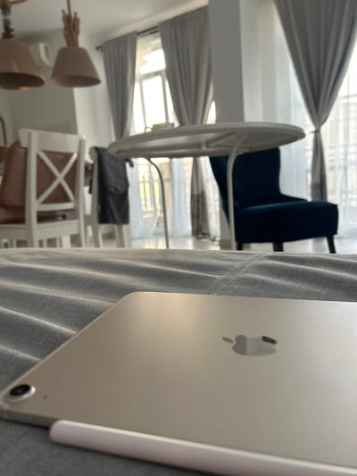 In a room in Chernomorets, Bulgaria, a tablet rests on a grey, textured surface in the foreground. The tablet is light-colored and features the Apple logo. An Apple Pencil is positioned beside it. In the midground, a white, round table with thin legs is visible. Behind the table, a dark teal armchair is placed near a window with grey curtains. Further back, two white chairs with brown seats and a black garment hanging on one of them are present. Two pendant lights hang from the ceiling. The background shows a window with light filtering through, suggesting it is daytime. The scene appears to be a residential interior, possibly a living room or dining area.