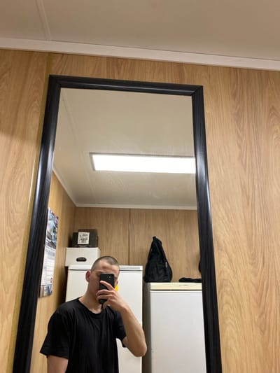 The image captures a mirror selfie of a male individual within an indoor setting in Марфино, Russia. The male, with a closely shaved head and wearing a black t-shirt, stands facing a large, dark-framed rectangular mirror. He holds a dark-colored smartphone with his right hand, obscuring the lower half of his face as he photographs his reflection. The visible background within the mirror shows light-brown wood-paneled walls and a white ceiling. A single, rectangular fluorescent light fixture is illuminated on the ceiling. To the left of the male's reflection, a calendar or poster hangs on the wall, beneath which a stack of white appliances, possibly refrigerators or freezers, is visible. On top of the upper appliance, a brown box and a white box labeled "DOKA" are present. To the right of the male's reflection, a black backpack rests on another white appliance. The scene depicts a functional, utilitarian room.