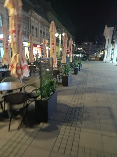 The image shows an outdoor seating area of a cafe at night, likely in Niš, Serbia, based on its appearance. In the foreground, there are several tables and chairs, with some of the chairs obscured by planters containing green foliage. Large, closed umbrellas, presumably for shade, are also visible, some with red and white branding. In the background, there are buildings with storefronts, illuminated by interior lights and street lamps. The street itself is paved with rectangular tiles, and shadows from the umbrellas and buildings are cast across it. A few people can be seen in the distance, walking along the street or in front of the shops. The overall atmosphere is quiet and dimly lit, typical of an urban setting at night.