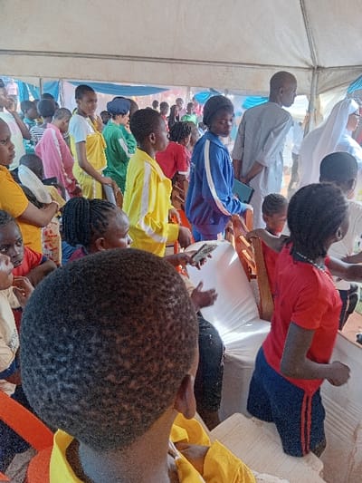 The scene depicts a crowded indoor-outdoor gathering under a large white canopy tent in Tuaniako, Nigeria. The majority of individuals are children and young adults of African descent, with a few adults interspersed. Attire is diverse, featuring several children in school-like uniforms, including yellow tracksuits with white stripes, blue tracksuits with white stripes, red t-shirts, and pink tops. One adult female is visible in a white religious habit, and an adult male wears a light-colored traditional garment. Most participants are standing and generally facing forward, observing an unseen focal point. Several individuals are holding or looking at mobile phones. Wooden chairs, covered in white fabric, are visible, some partially obscured by people. The environment suggests a daytime event, possibly a community or school function.