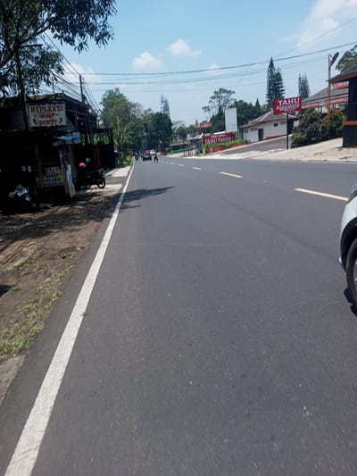 The image depicts a straight, asphalt road with a white line marking the edge and dashed yellow lines in the center. Buildings and trees line both sides of the road. Signage includes "REPLEKSI SEHAT" and "TAHU Sumedang." Motorbikes are parked on the left side of the road. A car is partially visible on the right. The scene appears to be a roadside area, with vegetation and structures alongside the pavement. There are a few distant figures walking on the roadside. The sky is blue with scattered clouds. The location is undetermined.
