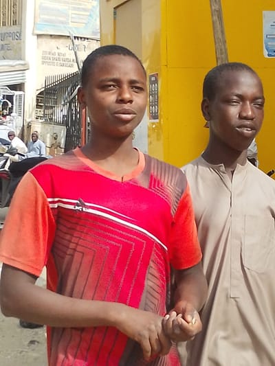 This image depicts an outdoor urban street scene in Babban Giji, Nigeria, under bright daylight. In the foreground, two young males of dark complexion are positioned. The male on the left wears a red short-sleeved V-neck t-shirt featuring an Adidas logo, white horizontal stripes, and a geometric pattern of darker lines over red and brown hues. His hands are clasped together at waist level, holding small, light-colored particles. He looks slightly to the right of the camera. The male on the right wears a light brown, buttoned-neck traditional long-sleeved garment and also looks slightly to the right, with his mouth slightly ajar.

The background on the left reveals a light-colored building with a partially visible blue sign displaying "PURPOSE UNITED". Below this, a black metal gate or fence features a white sign with black text indicating an address: "200 ROAD NO 10", "GIDAN SHARU GARBA", and "OPPOSITE GIDAN ASARE". Behind the fence, a motorcycle and two adult males in light-colored traditional attire are visible. The background on the right is dominated by a bright yellow wall or structure, adorned with a poster featuring a black grid pattern. A dark vertical post is also present on the right side. The overall scene suggests a commercial or residential street environment.