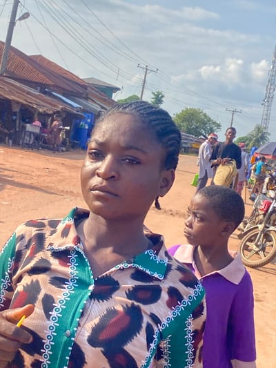 This image depicts an outdoor street scene in Amaseri, Nigeria, under bright daylight with a light blue sky and scattered white clouds. The ground is unpaved, consisting of reddish-brown dirt.
In the immediate foreground, a dark-skinned woman, appearing to be in her 20s or 30s, is positioned to the left. She has braided hair visible on her temple and wears a collared shirt. The shirt features a green placket and collar patterned with white circular motifs, contrasting with a body featuring a leopard print design in tan, dark brown, red, and black. Her expression is serious or contemplative, and she looks slightly to her left. A small yellow object is visible in her right hand. To her right and slightly behind her, a dark-skinned child, estimated to be between 5 and 10 years old, is partially visible. The child wears a purple long-sleeved shirt with a light-colored collar and looks towards the right.
The background reveals a street market environment. On the left, several structures with rusted corrugated metal roofs line the road, appearing to be market stalls or small shops. Blue plastic barrels are visible adjacent to these structures. Further down the street, multiple individuals are present; notable figures include a man in a black shirt holding a tan bag, and another man in a white shirt with a red headband. Overhead, numerous power lines extend from wooden utility poles, one of which supports a street light on the left. On the far right, a tall metal lattice tower stands. Parts of a parked motorcycle, including a white wheel, are visible behind the child, and an open umbrella suggesting a market stall is present on the right side. Green foliage and trees are visible throughout the mid-ground and background.