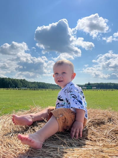 A young Caucasian child, appearing to be a toddler, is seated on a bale of hay outdoors. The child has light-colored hair and blue eyes and is wearing a white t-shirt with blue and black abstract designs and brown shorts with tiger-like patterns. The child is positioned on the right side of the frame, facing left and looking towards the camera with a smile. Their legs and feet are visible, with the feet bare.

The background consists of a vast green field under a bright blue sky with scattered white cumulus clouds. In the distance, a line of green trees borders the field, and a small, dark green building with a pointed roof is visible among the trees. The hay bale is textured and appears dry, with loose strands extending from it. The lighting suggests it is a sunny day.

The specific location is described as the city of Akhlebaikha (Kovriginskii S/S), Russia.