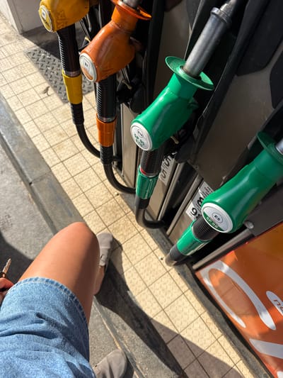 The image captures a view of a gas station pump with multiple nozzles, each colored differently to indicate the type of fuel. Green nozzles are present, with labels E5 SP 98 and E10 SP 95 E10 visible. Orange and yellow nozzles are also visible. The image appears to be taken outdoors on a sunny day. To the right, part of a person's leg, wearing a denim skirt and suede shoes, is visible. A set of keys is held in the hand. The location is Lançon-Provence, France.