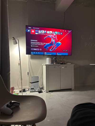 The image captures an indoor entertainment setup, viewed from an angled and rotated perspective. A large flat-screen television, mounted on a wall, displays the user interface of a PlayStation console. The screen prominently features "MARVEL Человек-Паук. Обновленная версия" (MARVEL Spider-Man. Updated version) with the character Spider-Man on a red background. The time "22:33" is visible in the top right corner of the TV.
Beneath the TV, a white cabinet holds a PlayStation 5 console, along with various wires and accessories, including a decorative metallic sculpture. A white floor lamp stands in the foreground, providing illumination. To the right, partially visible, is a chair or sofa armrest on which white PlayStation VR2 Sense controllers are resting. The floor is covered with light-colored, possibly marble-patterned, tiles.
Visible text on the TV screen includes menu options such as "Игры" (Games), "Мультимедиа" (Multimedia), "Продолжайте там, где вы ос..." (Continue where you le...), "Текущее событие: Главное событие" (Current event: Main event), "Показать подсказки" (Show tips), "Играть" (Play), "Призы" (Prizes), "Лоузья, которые играют" (Friends who play), "Справка по игре" (Game help), "111%", and "15+". On the ceiling, embossed letters spell "С Д" and "РОЖ", suggesting a "Happy Birthday" message (С Днём Рождения). The scene depicts a late-evening gaming session in a modern living space.