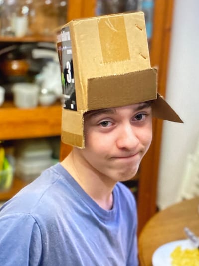 A young boy is wearing a homemade hat made from a cardboard box. The boy has light skin, green eyes, and short, dark hair. He is wearing a blue t-shirt and is looking at the camera with a slight smile. The background is blurred but appears to be an indoor setting with shelves and dishes visible. There is no identifiable information to place this image in Purkersdorf, Austria.