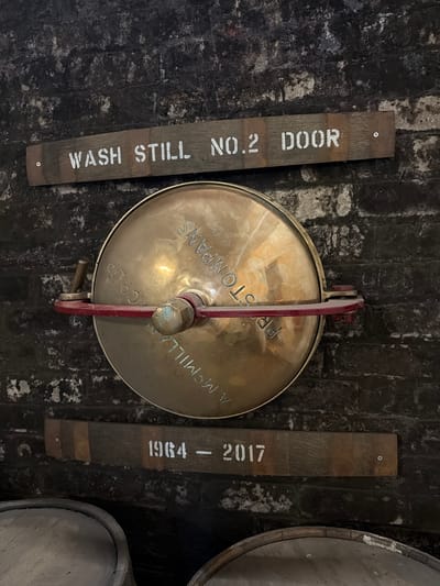 The image shows a close-up view of a dark, possibly aged wall in what appears to be a distillery or warehouse. The wall's surface is dark gray or black, with a rough, textured appearance possibly made of brick or stone.

Centered in the frame is a circular, metallic hatch or access port. It is held in place by a red metal bar that secures it tightly. Inscribed on the hatch are the words "PRESTONPANS A. MCMILLAN & CO LTD".

On either side of the hatch are two wooden planks. The plank on the left reads "WASH STILL NO.2 DOOR", and the plank on the right reads "1964-2017". The lettering on both planks is in white.

Fragments of two wooden barrels are visible in the upper-right and lower-right corners, suggesting that the location contains or is related to barrels of some kind.

Given the text on the hatch, the context of the room is likely within or near Blackford, United Kingdom, the known location of Prestonpans A. McMillan & Co Ltd. The presence of barrel fragments, the wash still reference, and the planks of wood suggest the interior of a distillery or maturation warehouse.