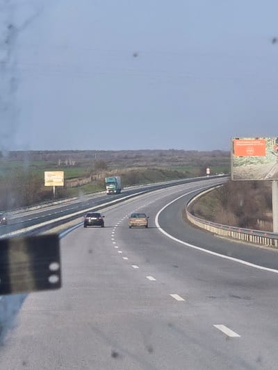 The image captures a multi-lane highway scene, located near Rupkite, Bulgaria, under an overcast sky. The perspective is from within a vehicle, with a dark, blurry object featuring two prominent white circular elements obscuring the lower left foreground, indicative of an interior component or window obstruction. Three vehicles are observed traveling away from the viewer: a light green semi-trailer truck in the leftmost lane, a dark-colored sedan in the middle lane, and a light brown sedan in the rightmost lane. A fourth lighter-colored vehicle is partially visible further down the road on the far right. The asphalt road features clear white dashed and solid lane markings. Metal guardrails line the road on both sides. On the right, a large billboard displays an orange background with Cyrillic text and an emblem, featuring an image of a person holding an axe. A smaller, light-colored sign is present on the left side of the highway. The surrounding environment consists of green and brown scrubland vegetation, transitioning to a distant treeline at the horizon. Utility lines are visible against the sky in the upper left. The image itself exhibits multiple dark, circular dust spots across the frame.