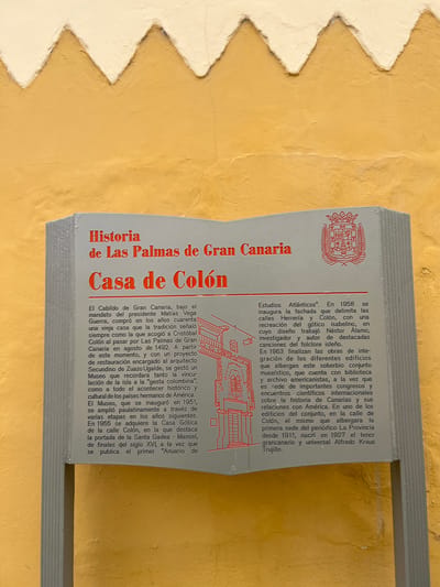 The image shows a placard in Las Palmas de Gran Canaria, Spain, detailing the history of the Casa de Colón (Columbus House). The sign features red lettering against a white background. The text explains the origins of the house as a place where Christopher Columbus stayed on his way to the Americas in 1492. It describes its transformation into a museum dedicated to the history and culture of the Americas and its relationship with the Canary Islands. The text highlights the architectural aspects, including the incorporation of the Casa Gótica and the gótico isabelino style facade. The environment is a wall.