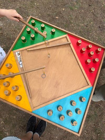 An overhead, eye-level shot captures a wooden ring toss game set up outdoors on a grassy, dirt surface. The game board is divided into four colored triangular sections: green, red, yellow, and blue. Each section contains several wooden pegs, some of which have rings on them. Two wooden sticks with strings and rings attached are positioned over the board, suggesting active play. A person's hand, holding one of the sticks, is visible in the upper left corner. The ground is covered with dry grass and dirt, indicative of an outdoor environment. The presence of a white sticker with Cyrillic text and numbers on the yellow section of the game board suggests a potential Belarusian origin, aligning with the specified location in Ракітніцкі сельскі Савет, Belarus.