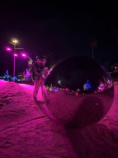 An outdoor night scene in Baan Lipa Noi, Thailand, is illuminated by vibrant pink and purple lights. The foreground is dominated by a large, highly reflective, spherical object, possibly a sculpture, set against a textured, pink-hued ground that appears to be sand or a similar granular material. To the left of the sphere, a person dressed in a silver, reflective astronaut-like suit stands. In the background, more abstract sculptures and stylized light fixtures are visible, including a lamppost with multiple lights. Palm trees can be seen silhouetted against the dark sky. Reflections of the surrounding lights and figures are visible on the surface of the large sphere.