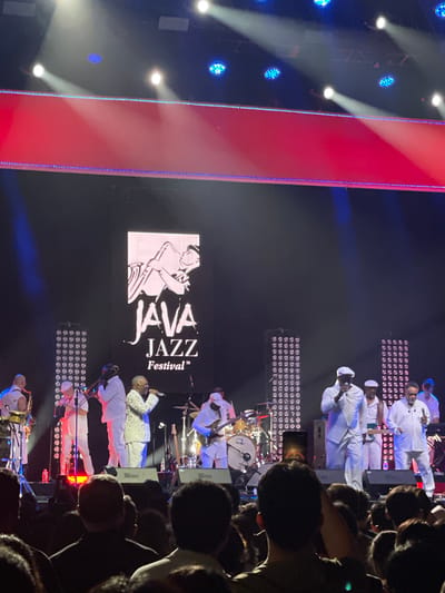 The image shows a live music performance at the Java Jazz Festival in Salembaran, Indonesia. The audience is visible on the right, with heads and shoulders of people densely packed. On the stage, a band dressed in white uniforms is performing. A screen behind the performers displays the Java Jazz Festival logo. There are stage lights above and behind the band. A large red light panel is seen on the left side of the stage.
