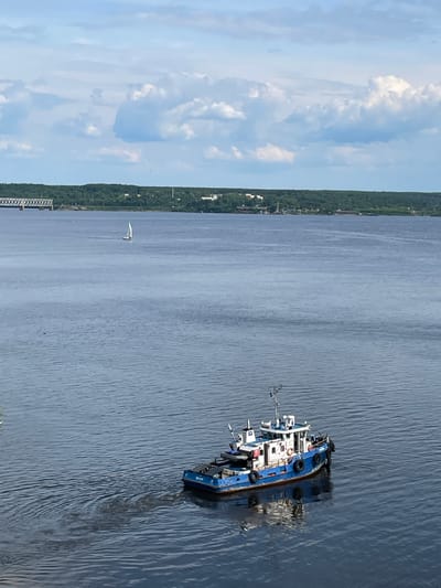 The image captures a wide expanse of water, likely a river, under a bright, partly cloudy sky. In the foreground to the right, a blue and white boat, resembling a tugboat or workboat, is prominently visible. It appears to be listing significantly or partially submerged, with its stern notably lower in the water, creating a pattern of ripples and disturbed water around it. Several black tires are visible hanging along its side, serving as fenders. Faint text, possibly "8160", is discernible on the stern of this blue boat.

Further in the distance, a smaller white sailboat with a single sail is seen gracefully gliding on the water. The far bank of the river is lined with a continuous stretch of lush green trees, indicating a forested area, interspersed with a few scattered buildings or structures. On the left side of the frame, close to the distant bank, a section of what appears to be a pier or a bridge structure is visible. The scene depicts a calm daytime setting, likely late morning or afternoon, in Пятый километр, Russia, with the listing blue boat being the central point of interest.