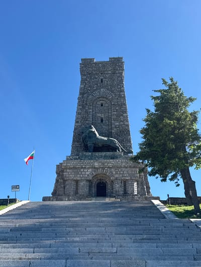 In the city of Shipka, Bulgaria, the Shipka Memorial is seen under a clear blue sky. A grand stone monument, shaped like a tower, dominates the center of the frame. Adorning its facade is a large, imposing bronze lion sculpture. To the left of the monument, the Bulgarian flag waves from a flagpole, its white, green, and red colors vivid against the sky. A tall, green fir tree stands to the right of the monument, its branches reaching towards the sky. In front of the monument, a wide, stone staircase ascends, leading up to the monument. The steps are made of large, gray stones. A ramp is also visible, providing an alternative access route to the side of the monument. In the distance, a small sign with arrows points the way to "Tickets." The scene appears to be a historical landmark, likely a memorial site, with a serene and solemn atmosphere.
