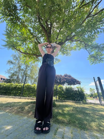 A young woman stands outdoors on a sunny day in Chotěšov, Czechia. She is positioned in the lower right portion of the frame, dressed in a black top and black wide-leg pants, with black sandals on her feet. She has her hands raised above her head, holding a clear plastic bottle. The camera is angled upwards, capturing a large tree with lush green leaves that dominates the upper left of the image. The sky is a clear blue. A metal fence and greenery are visible behind her, along with a patch of grass and a paved walkway. The scene suggests a casual outdoor setting, possibly a park or garden, under bright daylight. There is no visible text in the image.