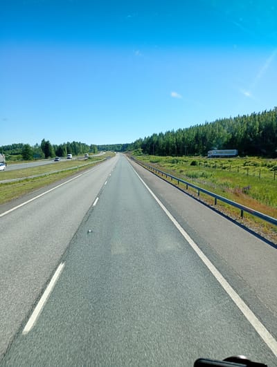 The media file shows a highway scene in daylight, likely during the summer due to the lush green vegetation and clear blue sky. The perspective suggests the photo was taken from a vehicle moving along the road. The highway itself is paved, with clear white lane markings. On the left side of the road, there is a grassy median and then another lane of traffic with a few cars visible. On the right side of the road, there is a metal guardrail separating the highway from a grassy field bordered by a dense forest of evergreen trees. The sky is bright blue with a few wispy clouds.

In the distance, on the right side of the highway, a white truck with text on its side is visible parked in the field. The text on the truck reads "SUOMEN PORAKAIVO". This suggests the location is in Finland, as "Suomen" means "Finland's" and "Porakaivo" means "drilling rig" or "well drilling." The overall scene is peaceful and serene, characteristic of a sunny day in a rural or semi-rural area. The time of day appears to be mid-morning or early afternoon given the bright, direct sunlight. There are no people prominently visible in the image.