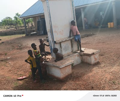 The photograph depicts a group of children in Gidan Kogi, Nigeria, standing and sitting around a weathered, light-colored structure with a large, upright panel. The structure appears to be composed of concrete blocks. The children are of varying sizes and wear different clothing, including a yellow and black shirt, a striped shirt, and a pink shirt. Some are bare-chested. The ground is a reddish-brown soil. In the background, a building with a corrugated metal roof supported by columns is visible. Individuals can be seen inside the building. Vegetation and other structures are sparsely distributed in the distance. A red plastic lid sits on the ground near the children. The scene is bathed in sunlight.