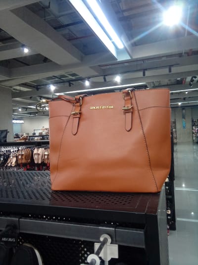 The image shows a brown leather Michael Kors tote bag displayed on a black metal shelf in a retail store. The store has overhead fluorescent lighting and appears to be a department store or shoe store, with rows of shoes and other merchandise visible in the background. The location is inferred to be in a retail environment, potentially within the city of Parroquia Guatire, Venezuela.
