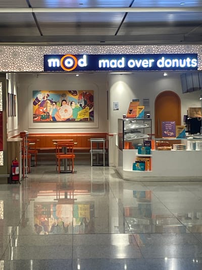 The image shows the exterior of a "Mad Over Donuts" shop. The shop has a bright neon sign with the logo "mod" and the words "mad over donuts" written in white. Below the sign is a colorful mural depicting people holding donuts and coffee. Orange banquettes and tables with chairs are visible in front of the mural, suggesting a seating area. A glass display case is filled with donuts and other items, and a counter is present. In the background, an arched orange doorway and various promotional posters can be seen. The floor is highly reflective, mirroring the shop's interior. To the left, a red fire extinguisher is mounted on a stand. The overall environment appears to be an indoor public space, possibly an airport or a mall, given the reflective flooring and ceiling design. There are no people clearly visible in the image, though figures are depicted in the mural. The location of Gollapalle Khurd, India, is not visually evident from the provided image.