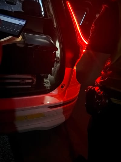 The image depicts a close-up, low-angle view inside the trunk of a white vehicle, illuminated by red ambient lighting, likely from the car's taillights. A tablet displaying a keyboard is visible on the left, positioned atop what appears to be a black case or container. Below this, another dark rectangular object is present. Further back and to the left, a silver, rectangular case with latches is visible. In the right portion of the frame, a person is partially visible, wearing a dark-colored uniform or jacket and holding what seems to be a tool or device. The exterior of the car's bumper is visible at the bottom, with parking sensors and a yellow stripe. The scene is dark, with the red lighting creating strong shadows and highlights. The overall impression is that of an operational setting, possibly related to law enforcement or emergency services, given the presence of a tablet and the uniform. The image was taken in the city of Smyadovo, Bulgaria.