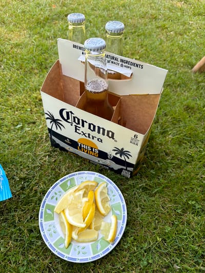 An overhead, eye-level medium shot captures a six-pack of Corona Extra beer bottles, one of which is open and half full, resting on lush green grass. The beer box, partially visible, displays the Corona Extra logo and a tropical scene with palm trees and a sun. Beside the box, a white plate with green and blue floral accents is laden with several lemon wedges. The scene appears to be set in an outdoor environment, likely a garden or park. The overall impression is one of a casual gathering or relaxation. There are no discernible landmarks or indications of the specific location within the city of Papenburg, Germany.