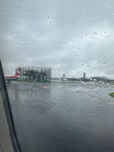 The image shows a view from inside an airplane window. The window is covered in rain droplets, obscuring the view of the tarmac. The tarmac is wet, and there is a small red-and-white airplane parked nearby. The image was taken at Narita International Airport in Narita, Japan (35.772, 140.391). The sky is grey and overcast.
