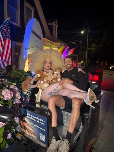In Provincetown, Massachusetts, at night, a drag performer with a large blonde wig and dramatic makeup sits in a float, holding several teddy bears. They are wearing a gold dress and have tattoos on their arms and legs. A man is seated next to the performer, with his arm around the performer's shoulder and his leg resting on the performer's lap. He is smiling and wearing a black shirt and shorts. The float is decorated with flowers and has a sign that reads "Make sexual health a part of your self-care. CareThatFitsYou.org." In the background, there is a building with an American flag and rainbow flags. The scene appears to be part of a parade or event.