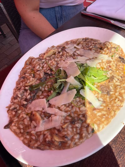 A high-angle, full-frame shot shows a plate of risotto on a dark, possibly wooden, surface. The risotto is garnished with arugula and shavings of parmesan cheese. A person's arm and leg are visible on the left side of the image, suggesting the photograph was taken by someone seated at the table. A napkin and a metal object, possibly cutlery, are also partially visible next to the arm. The location details (Guildford, UK) are not discernible from the image alone.