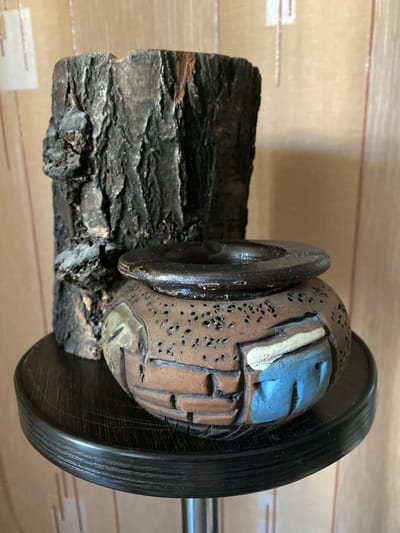 A rustic wooden piece, resembling a charred log, stands behind a small, earthy-toned ceramic pot with a textured surface and decorative elements. The pot features a dark rim and a design that incorporates brick-like patterns and a blue section with carved indentations. Both objects are positioned on a dark, circular wooden stand with grooved detailing. The background consists of a textured, light brown wall. The scene is indoors, likely a residential setting, and no specific location within Novaya Usman', Russia, can be determined from the visual information.