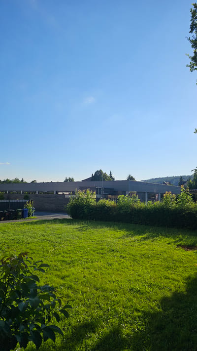 The image depicts a sunny day with a clear blue sky over a suburban or semi-rural area. In the foreground, a well-maintained green lawn is visible, with a shadow cast from the left side, indicating the sun's position. A low-lying bush with dark green leaves occupies the bottom left corner.
In the mid-ground, a row of manicured hedges forms a green barrier. Behind the hedges, several dark-colored trash bins are lined up against a fence. Further back, a long, low-rise building with a grey facade and a dark roof stretches across the scene. The building appears to be a commercial or institutional structure, possibly a school or office building, given its size and simple design. Trees with green foliage are scattered behind the building and on the hills in the distance, suggesting a landscape with natural elements.
The overall impression is one of a peaceful, well-kept environment, likely an area adjacent to a public facility or residential zone. The bright sunlight and clear sky contribute to a sense of warmth and clarity. The location is identified as Hann. Münden, Germany, a town known for its historic architecture and scenic surroundings, though this specific scene appears to be a more modern development within the area. There are no people or active events captured in the image.