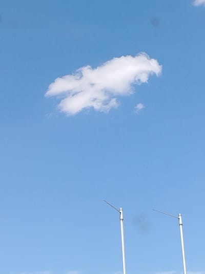 This is a photograph of the sky in Kirchardt, Germany. A single, white cloud with an unusual, perhaps anthropomorphic, shape is visible in the upper portion of the frame. The cloud appears to have two distinct "legs" extending downwards and a rounded "body" above. The sky is a clear, pale blue, indicating daylight. In the lower half of the image, two tall, slender poles with horizontal attachments are visible, positioned apart from each other. These poles are metallic and appear to be part of some outdoor infrastructure, possibly streetlights or antenna supports. A faint, dark smudge, possibly a distant object or lens artifact, is present in the sky between the two poles.