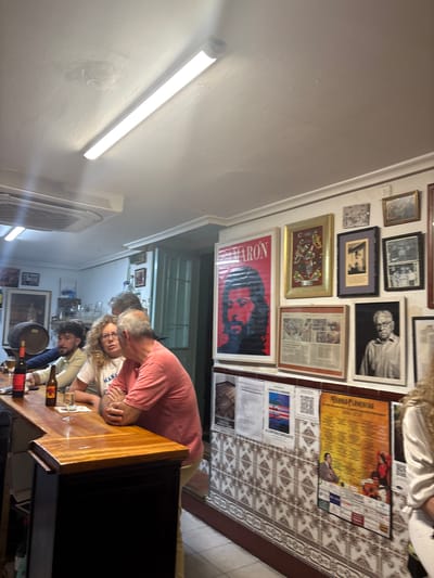 The image shows the interior of a bar or restaurant in Córdoba, Spain. Several people are gathered at a wooden bar. A man in a salmon-colored shirt is leaning over the bar, possibly talking to a woman. Other people are seated at the bar, drinking, and conversing. Various framed photographs, posters, and prints adorn the walls, creating a dense gallery-like display. A long fluorescent light fixture is visible on the ceiling, providing illumination. The floor has decorative tiles. Bottles, glasses, and other bar paraphernalia are visible on the bar surface.