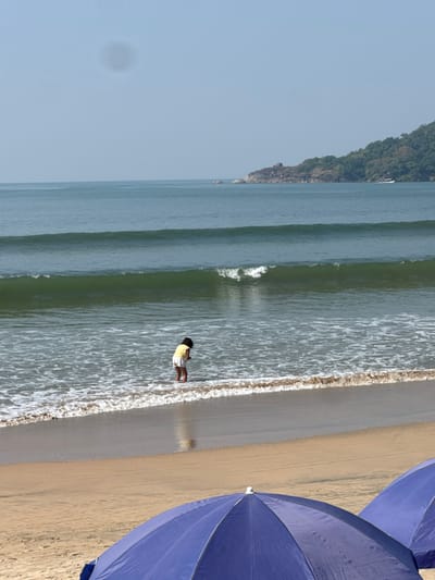 A beach landscape featuring calm ocean waters meeting a sandy shore. A person stands at the edge of the water, clad in a yellow top and shorts. To the right, the vibrant blue of beach umbrellas dominates. A small, tree-covered landmass juts into the sea in the distance, under a clear sky. The image is horizontally oriented, emphasizing the long stretch of beach and sea.
