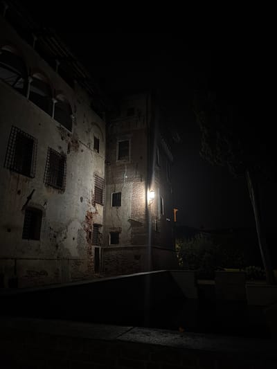 An outdoor night scene in Lagnasco, Italy, features an aged building with weathered walls and barred windows. The building, constructed of brick and plaster, exhibits significant signs of decay, with portions of the facade crumbling to reveal underlying brickwork. A streetlamp affixed to the building illuminates its facade with a warm glow. Adjacent to the building, a dark, unlit area is visible, with hints of greenery and a tall, dark tree silhouette against the night sky. The overall atmosphere is one of historical neglect and somber tranquility.