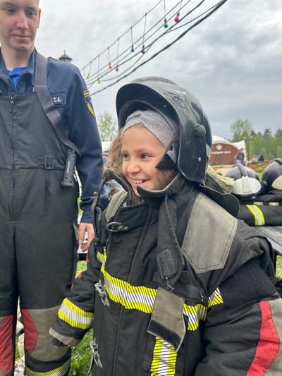 A young child wearing a black firefighter's helmet and a firefighter's coat poses with a man. The child is smiling and looking down and to the right. The firefighter's coat is black with yellow reflective stripes. The man in the photo is wearing a blue shirt and black overalls. "WORKLIGHT" is written on a cylindrical object clipped to his chest. The background of the photo includes green grass, trees, buildings, and lights strung on a wire. While the scene seems to be at an outdoor event, the location cannot be definitively confirmed as Grebnevo, Russia based on the provided image.