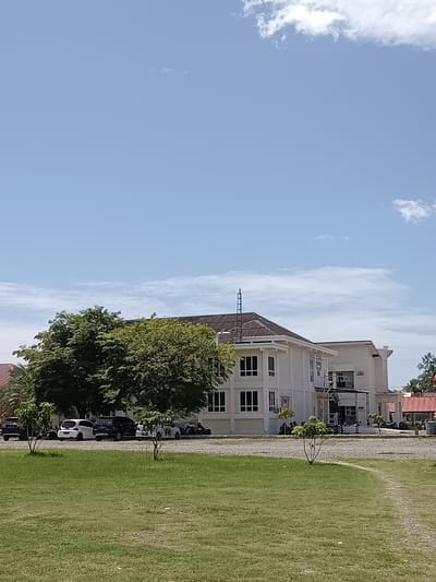 The image depicts a modern building with a white exterior and dark-framed windows, situated in a semi-urban or suburban area. The building appears to be a multi-story structure, with a darker-colored roofline. In front of the building, there is a gravel parking area with several vehicles, including cars and motorcycles. Adjacent to the parking area is a lush green lawn, with a pathway curving through it. Several trees are scattered throughout the scene, with large, leafy trees providing shade near the building and smaller, sapling-like trees in the foreground. The sky is a clear blue with scattered white clouds. Based on visual cues and the context of Mesjid Reuntoh, Indonesia, this scene likely represents a government building, office complex, or educational institution. There are no visible people in the image.