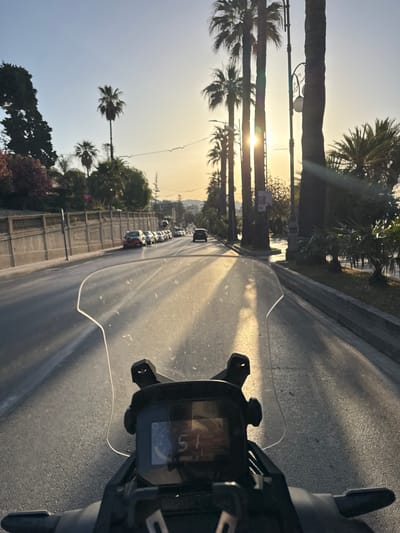 The image shows a point-of-view shot from a motorcycle on a paved road in Sanremo, Italy, at sunset. The road is lined with palm trees and a stone wall. Several cars are parked along the side of the road. The motorcycle's digital dashboard is visible in the foreground, displaying a speed of 51. The sun is low in the sky, casting long shadows and creating a golden light.