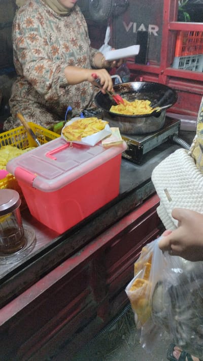 The image depicts a street food vendor in Sambungrejo, Indonesia, preparing food. A woman wearing a floral patterned tunic and a hijab is actively cooking in a large pan, using red tongs to handle the food. The pan sits on a portable stovetop. In front of the vendor, a bright pink plastic container is visible, topped with a serving of cooked food on white paper. To the left, a yellow basket contains more food items.
In the background, the word "VIAR" is visible on a red structure, possibly indicating a brand or the name of the establishment. The setting appears to be an outdoor market or roadside stall, suggested by the rustic counter and the dimly lit environment, hinting at either early morning or evening.
On the right side of the frame, a person's hand is shown holding a white woven bag and a plastic bag filled with food, indicating a customer's purchase. The overall scene conveys the hustle and bustle of a food vendor's daily operations, capturing a moment of food preparation and transaction in a local Indonesian setting. The focus is on the cooking process and the exchange between vendor and customer.