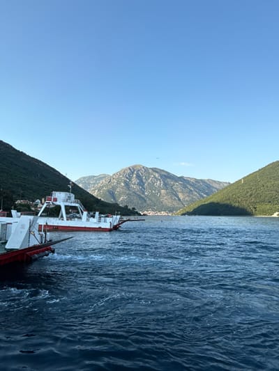 In Jošice, Montenegro, a ferryboat navigates the choppy, dark blue waters of a bay, bordered by steep, verdant mountains on either side. The ferry, predominantly white with red accents, has a multi-level superstructure and a distinctive red prow. The water is disturbed by the movement of the ferry, creating white foam and ripples. In the distance, a town with white buildings is nestled at the foot of the mountains, and further back, rocky peaks rise against a clear, pale blue sky. The overall scene captures a moment of transit within a stunning natural landscape.