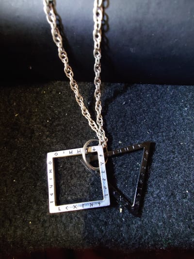 A close-up, eye-level shot captures a silver necklace with two pendants against a textured dark surface. The necklace has a fine, twisted chain. The left pendant is a silver square outline, with a darker silver inner border, inscribed with a series of small symbols that appear to be runes along its edges. The right pendant is a dark, possibly black metal, triangular outline. The overall lighting is dim, with a slight highlight on the chain and the silver pendant. The background consists of a dark, fuzzy material, likely fabric or felt, and a smoother, darker surface above. There are no people or animals visible. There is no indication of the city of Sokolohirsk, Ukraine in the image.