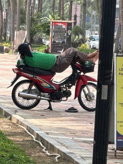 A person is observed lying prone on a red Honda Wave motorcycle, parked on a paved surface in An Hải Ward, Vietnam. The individual wears a green t-shirt, dark trousers, and a dark baseball cap, with their head resting on the rear seat. Their bare feet are elevated and positioned over the motorcycle's front handlebars, above a small black basket containing a green helmet. The motorcycle is supported by its kickstand. A pair of black sandals rests on the pavement near the front wheel. In the background, numerous palm trees line the area, with a red vending machine visible to the left of the frame. Further back, a white car and other motorcycles are partially visible, suggesting a road or parking area. A black utility pole and a yellow sign with Vietnamese text are present on the right side of the frame. A narrow strip of soil with some sparse vegetation and a white hose-like object runs parallel to a concrete curb in the foreground.