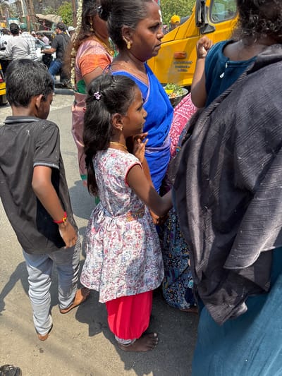 In a scene photographed in Ambernath, India, a group of people is gathered near a yellow auto-rickshaw, likely in a street setting. A woman in a blue sari and a girl in a white floral dress with a red underskirt are central figures. Another woman is wearing an orange sari. A young man in a black t-shirt and grey pants is also part of the group. Some individuals are barefoot. The presence of other people, a decorated garland and a city street indicates that this could be a public or religious event. There is an item with a yellow license plate present.